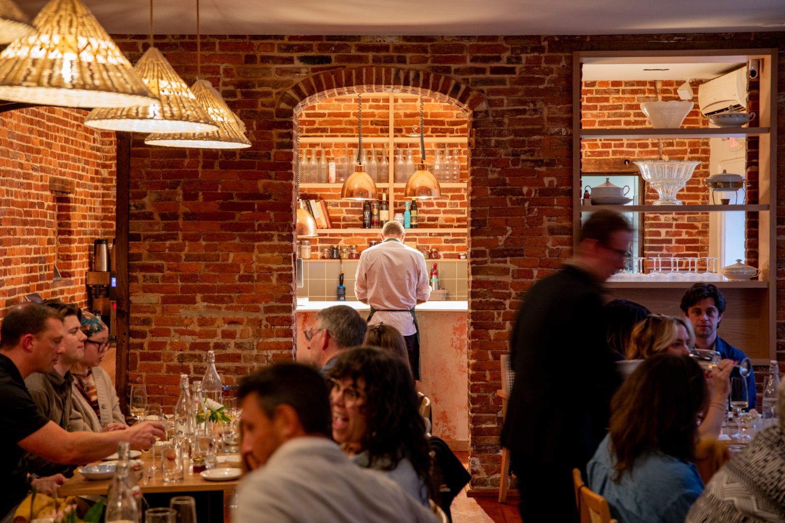 Cozy restaurant with warm lighting and exposed brick walls. Diners enjoy their meals while a chef works at an open kitchen. The atmosphere is lively and inviting.