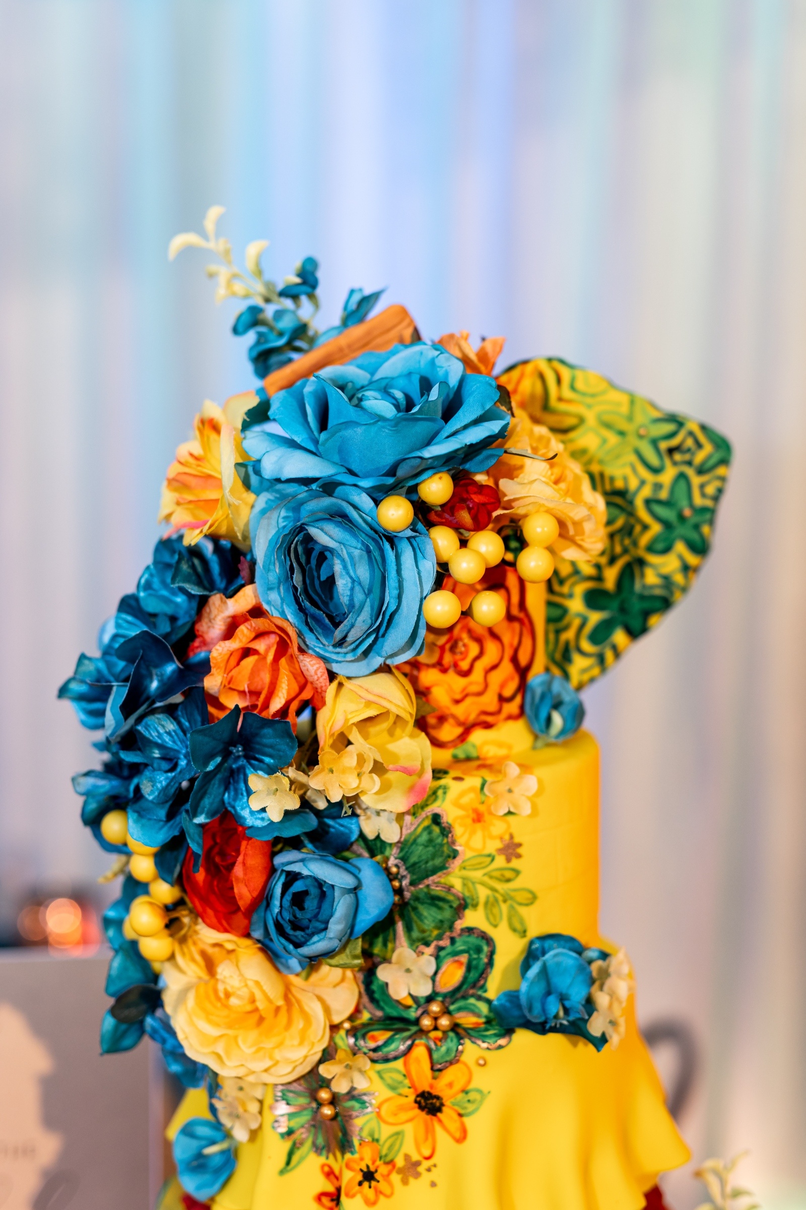 Close-up of a vibrant yellow wedding cake adorned with bold blue, orange, and yellow sugar flowers and intricate hand-painted floral designs.