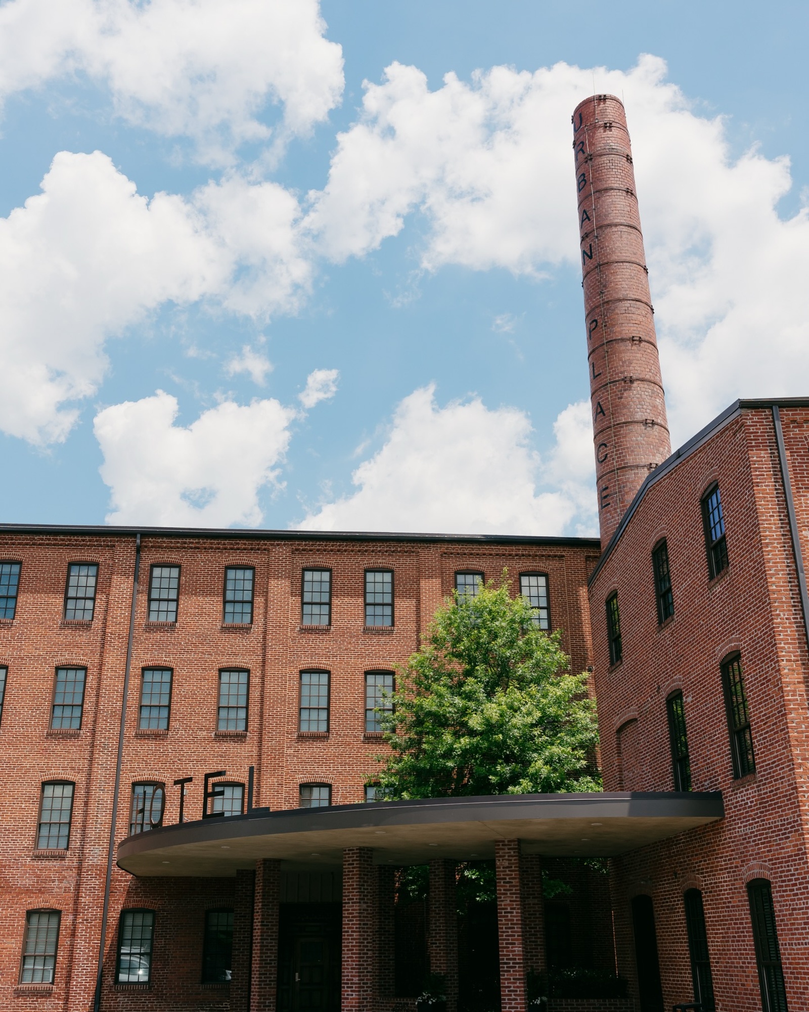 Historic red-brick boutique hotel with tall smokestack and black window frames under a bright blue sky with clouds.