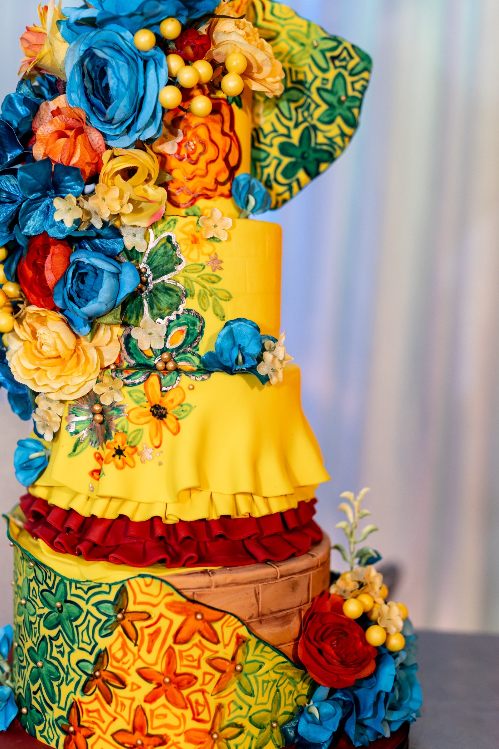 Three-tier yellow wedding cake decorated with colorful sugar flowers, red ruffles, and intricate green and orange patterns inspired by Latin design.
