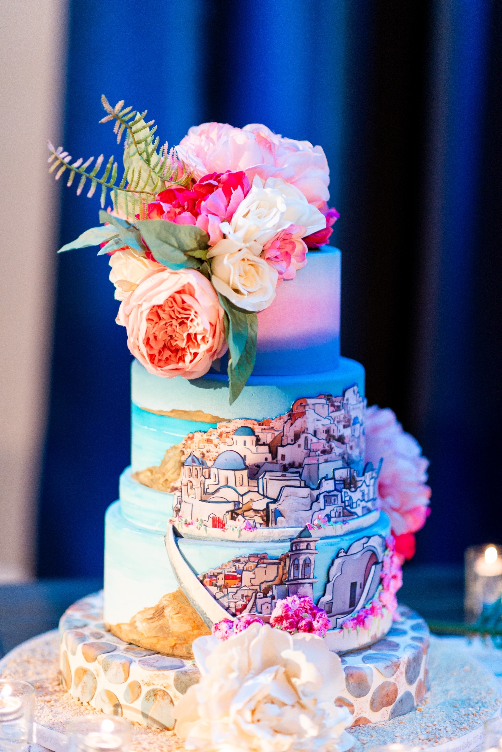A three-tier wedding cake decorated with hand-painted scenes of a Mediterranean village, featuring white and blue domed buildings resembling Santorini. The cake is topped with vibrant pink, peach, and white flowers, accented with greenery. The base tier is designed with a stone-like pattern, and the cake is displayed on a sandy surface surrounded by small candles.