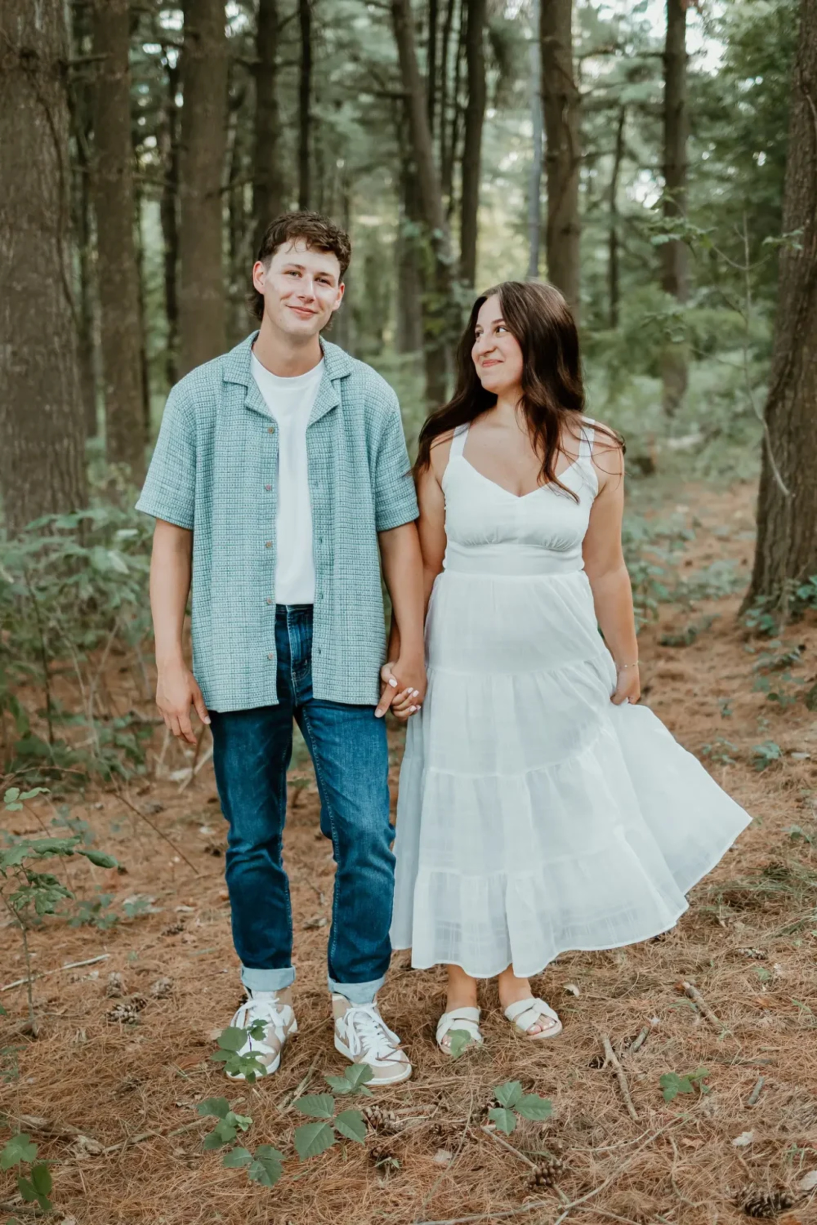 Couple holding hands and smiling among tall pine trees during their outdoor engagement shoot at Sam Lewis State Park in York County, Pennsylvania.
