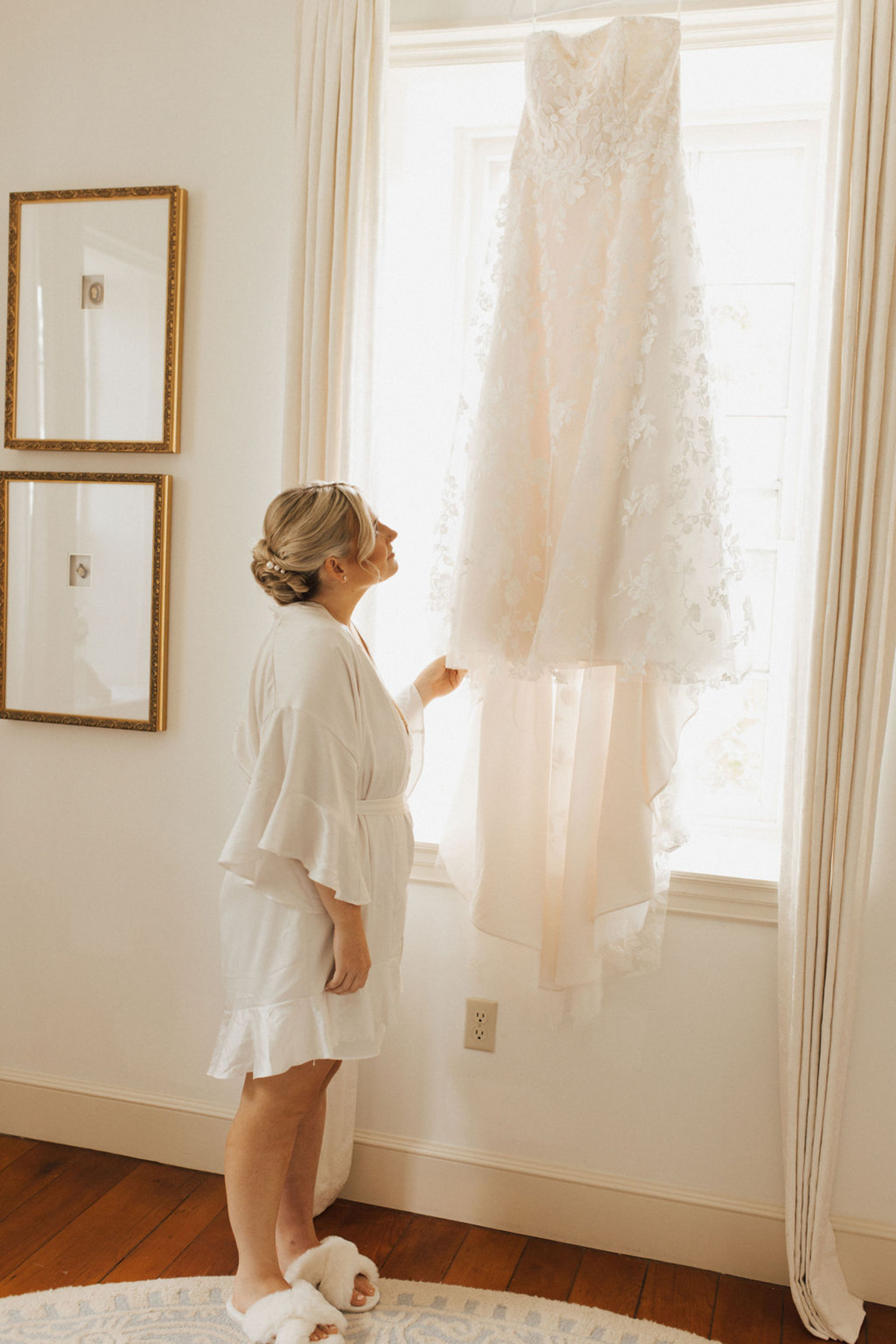 Central PA bride wearing white silk rob, white fuzzy slippers and low bun hairstyle admires lace strapless wedding dress hanging in front of window