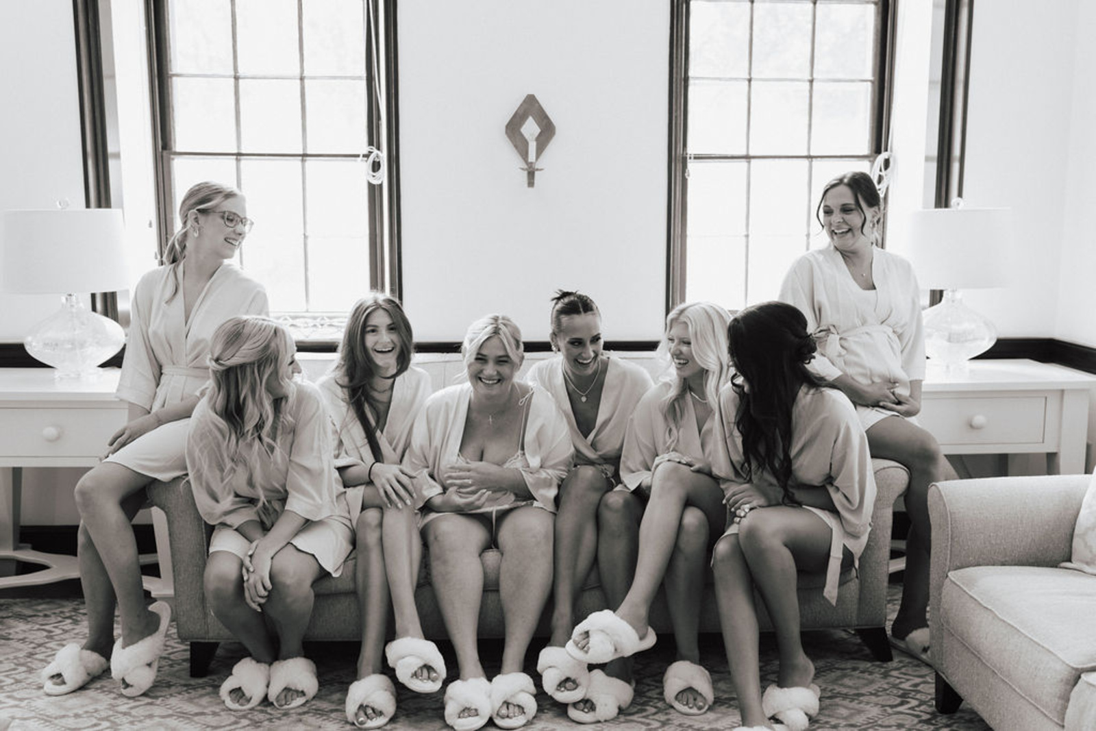 Black and white image of bride and bridesmaids smiling and sitting on couch in matching robes and fuzzy slippers