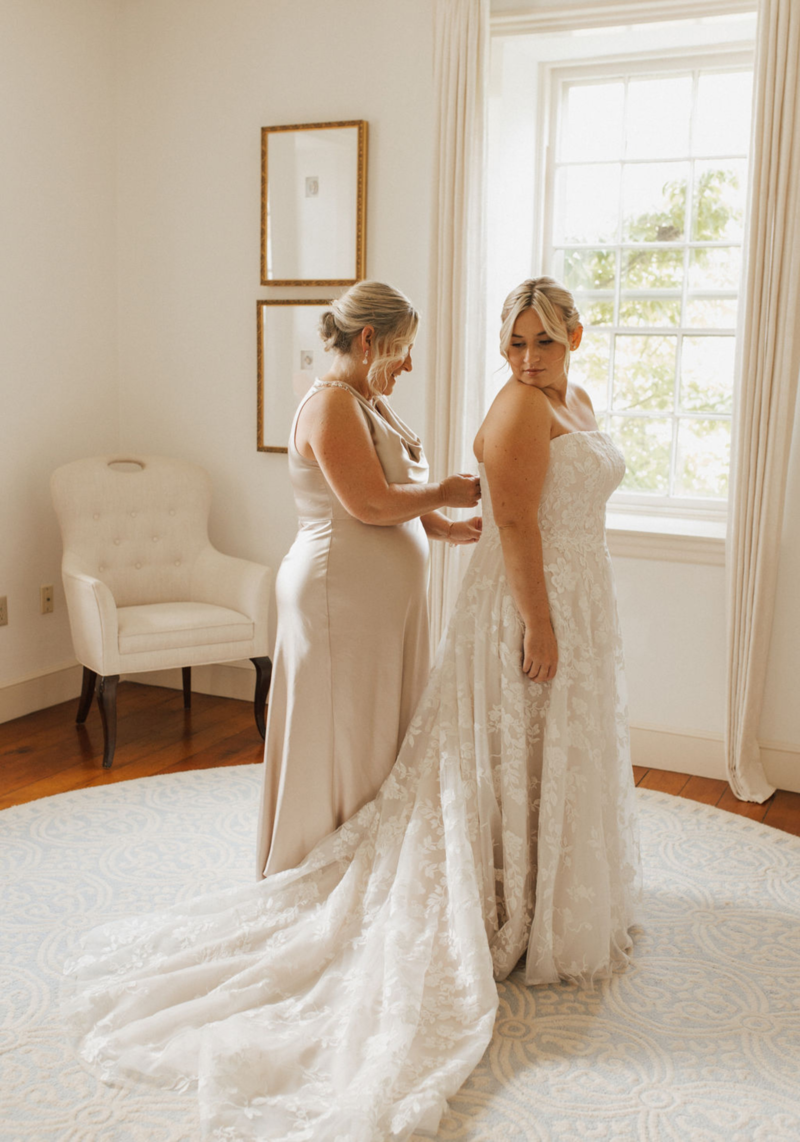 Central PA in a strapless lace wedding gown looks over her shoulder as the mother of the bride in a champagne silk long dress zips the bride's gown in the back
