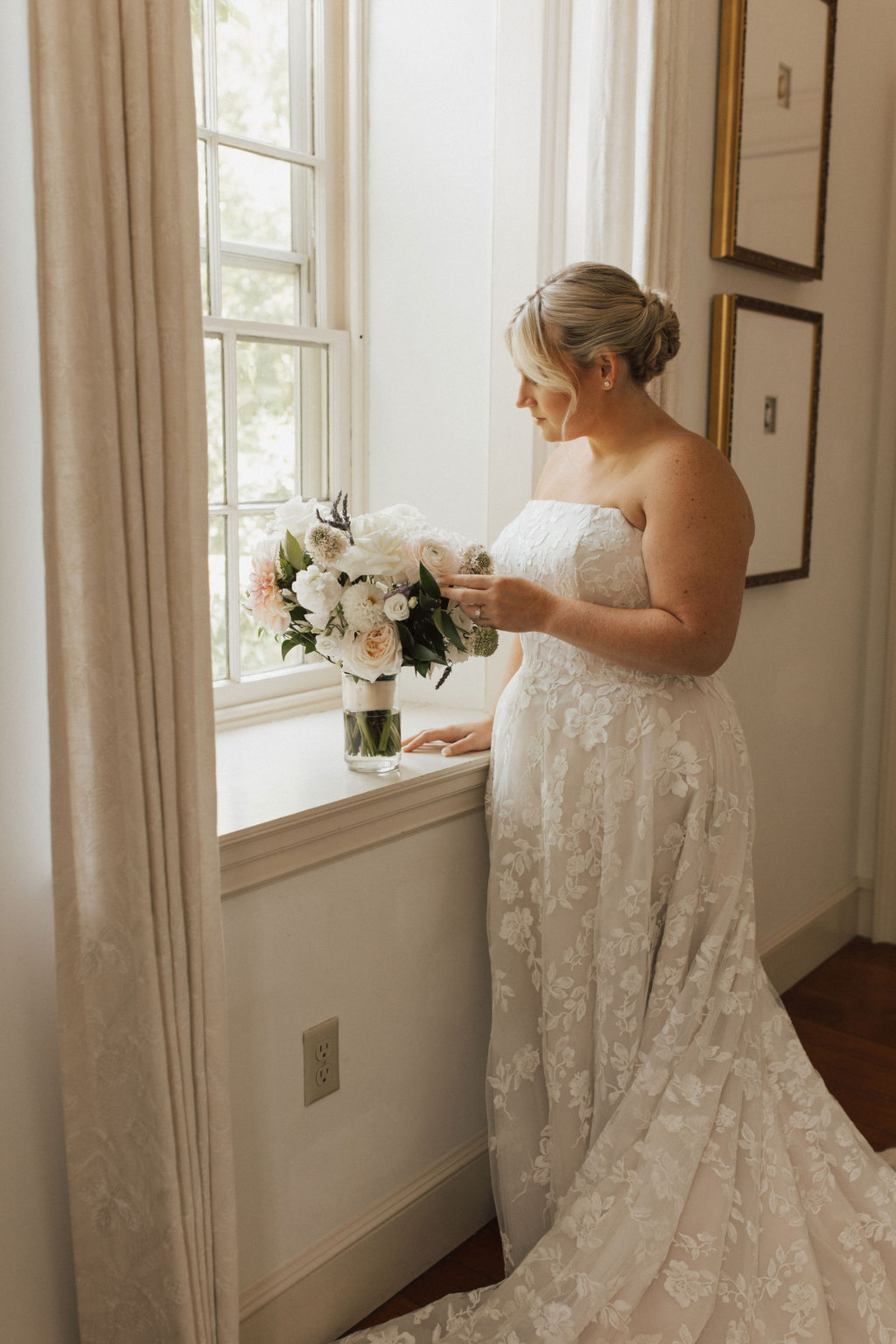 Central PA bride in a lace strapless wedding gown and low bun hairstyle admiring classic white and blush bouquet by the window