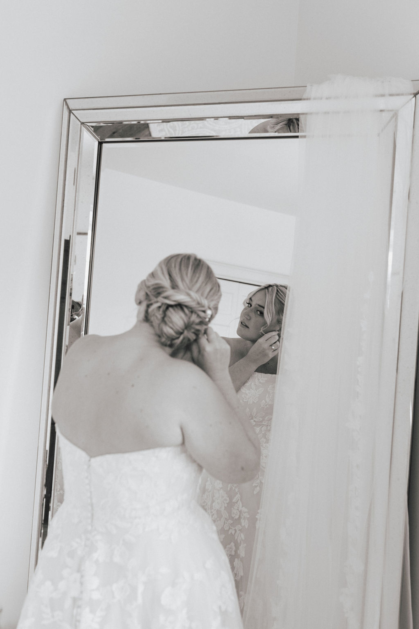 Black and white image of the back of the bride wearing a lace strapless dress looking in a mirror as she puts on her earrings