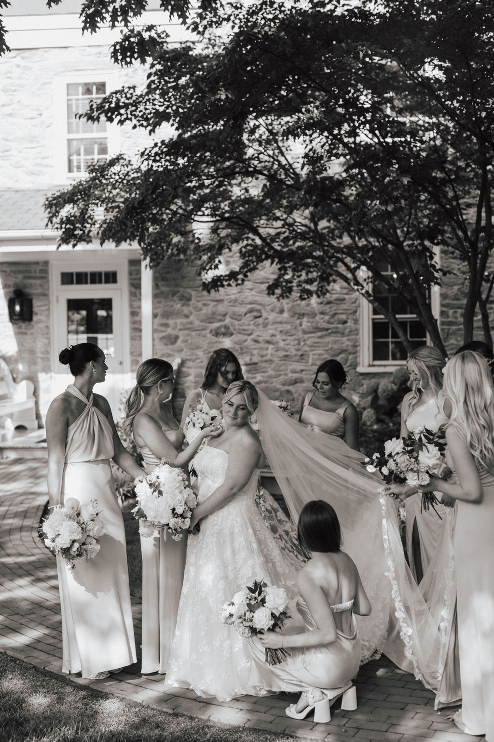 Black and white image of the bride in a lace dress and cathedral veil surrounded by 7 bridesmaids fixing her train and veil