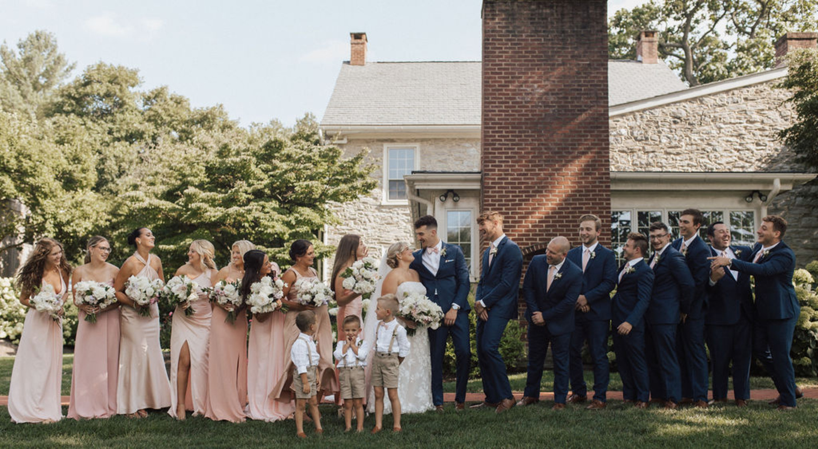 Bride in a strapless lace gown and groom in a navy suit surrounded by bridesmaids in long pale pink dress and groomsmen in navy blue suits