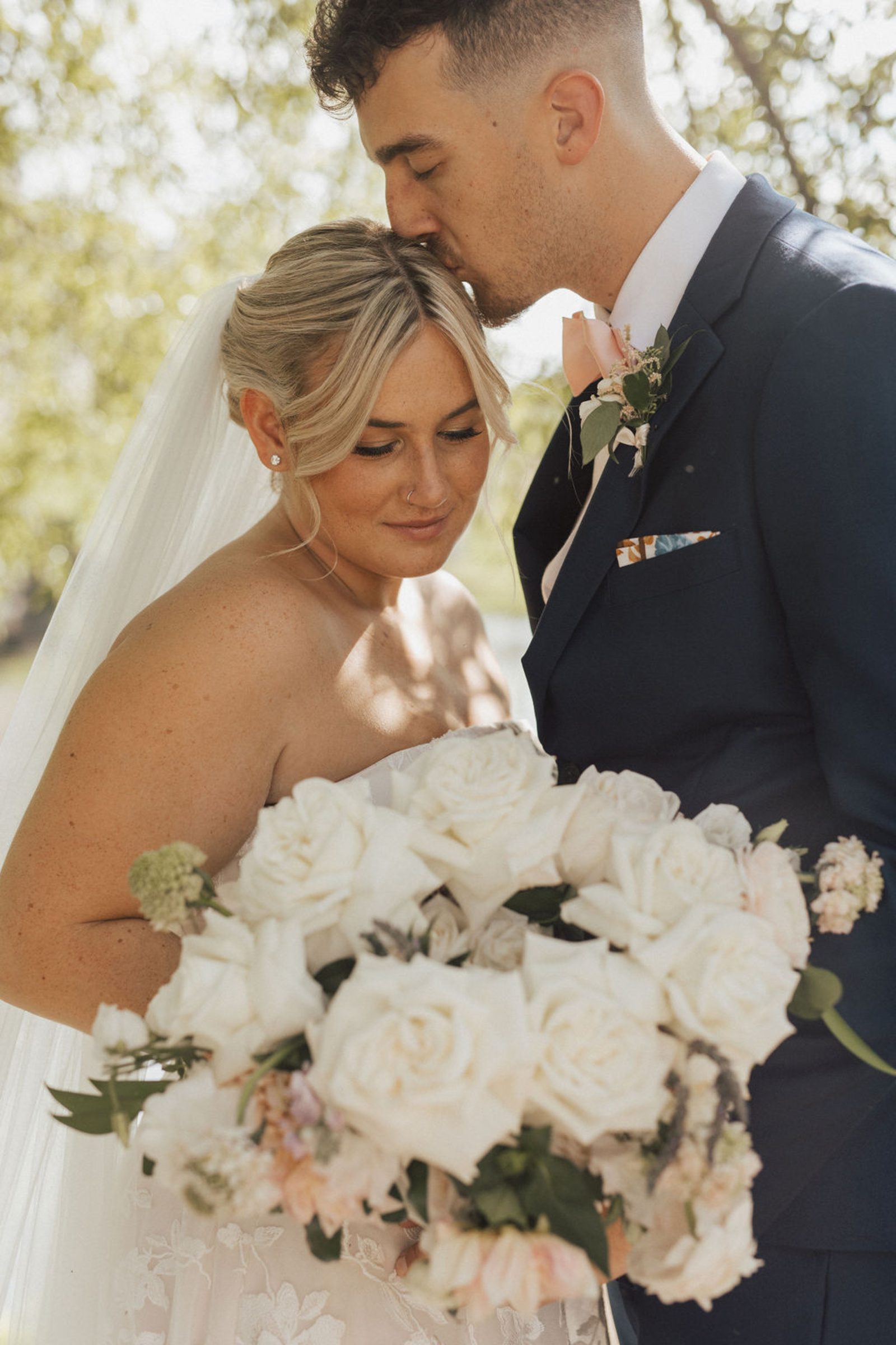 Central PA bride holding a beautiful white rose wedding bouquet while groom in navy blue suit kisses her on the top of her head