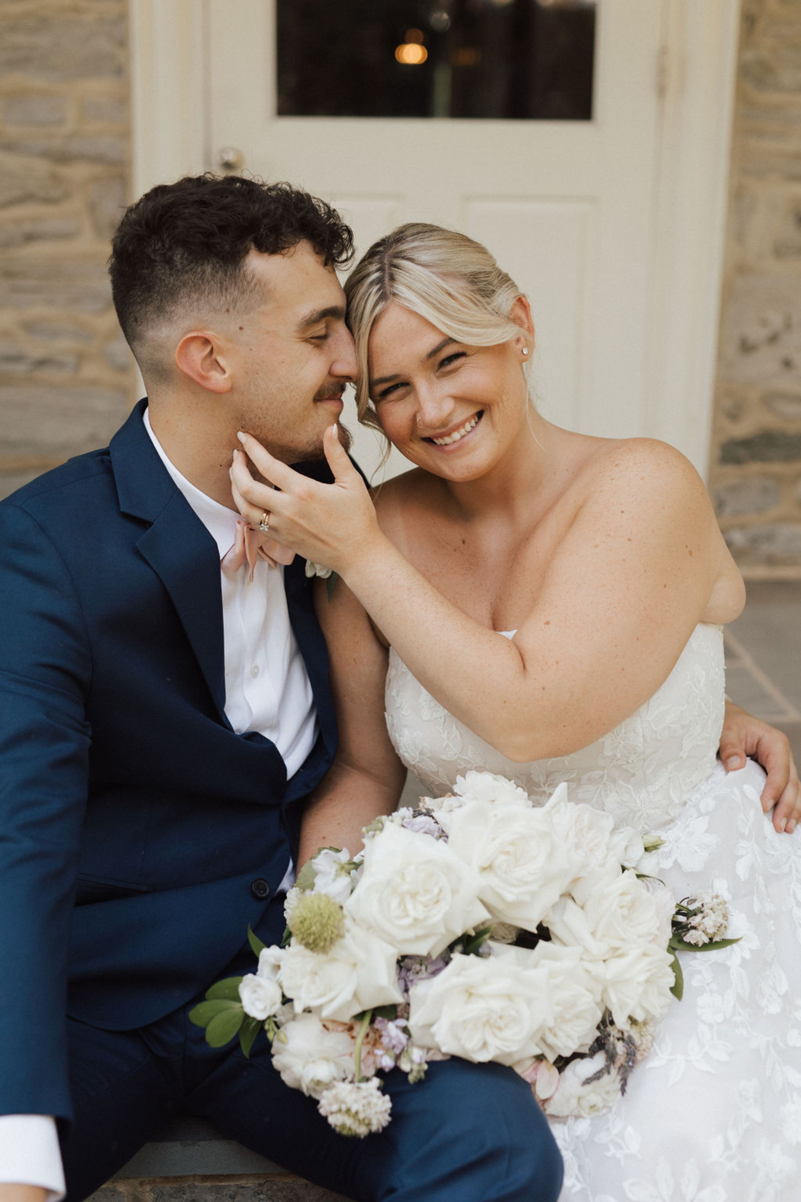 Central PA newlywed bride and groom sitting on steps in a navy suit and strapless lace dress smiling and holding white wedding bouquet