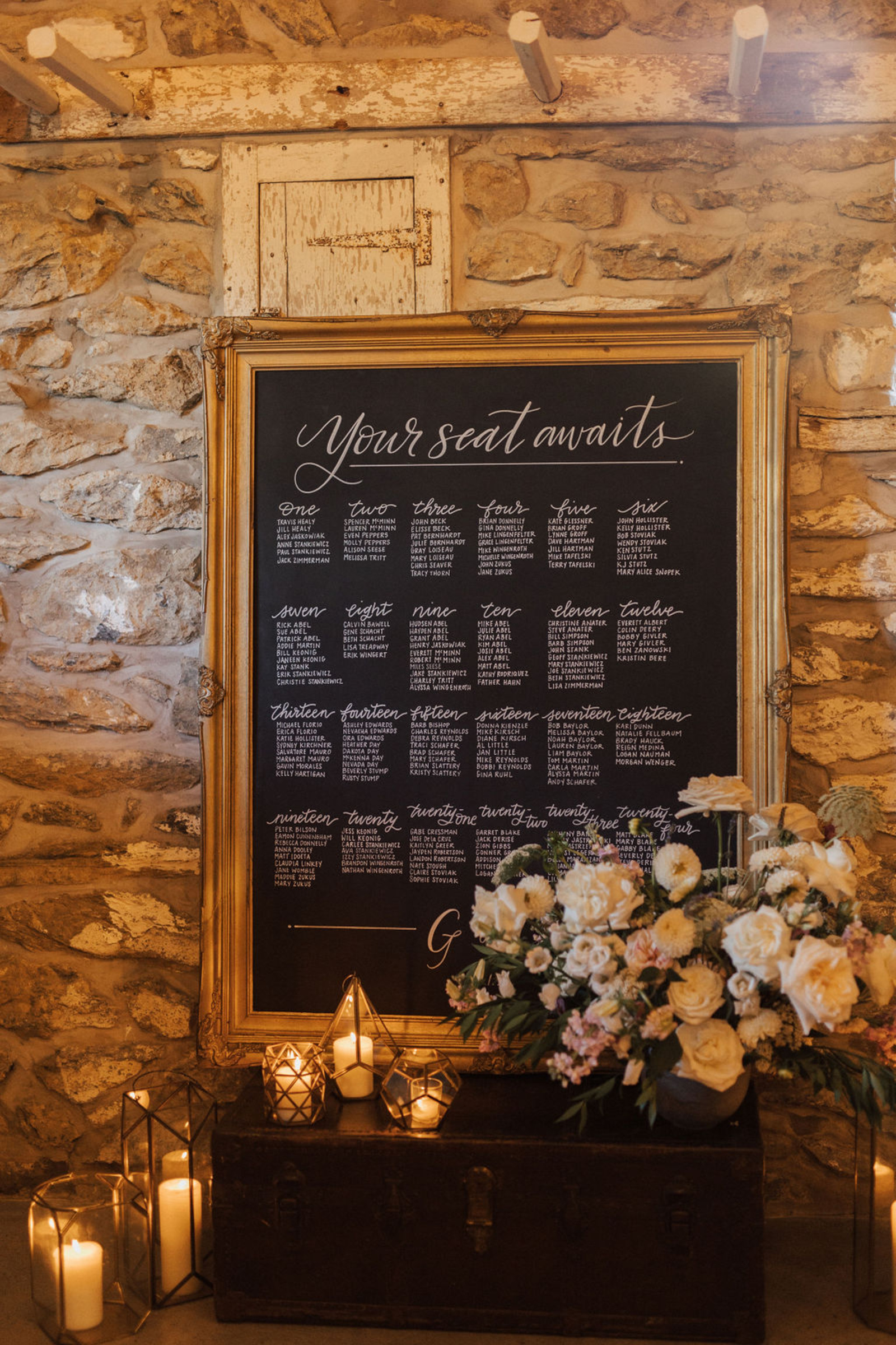 Large chalkboard with a thick, ornate gold frame used as a seating chart against a stone wall on top of an antique trunk surrounded by romantic candles and florals