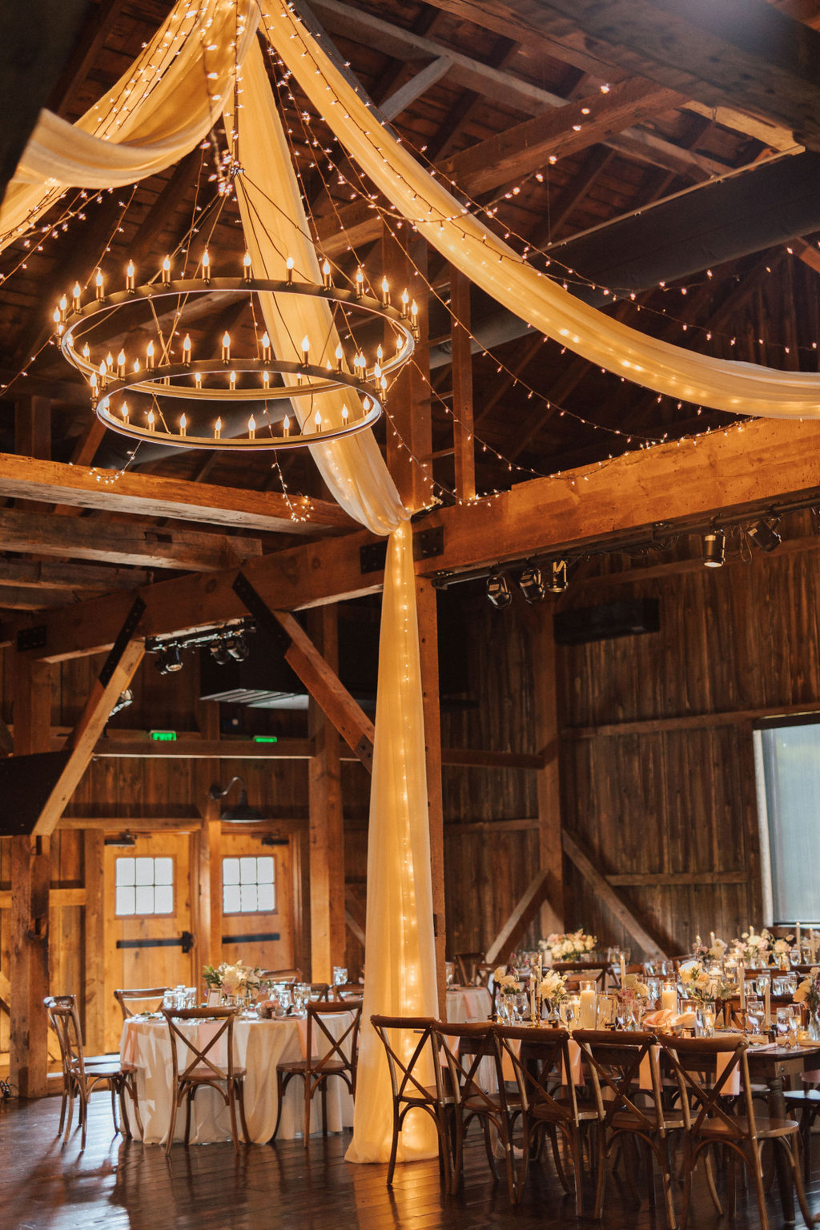 Rustic Central PA venue with wood beams and a large rustic two-tier chandelier with white drapery on the beams and strings of lights