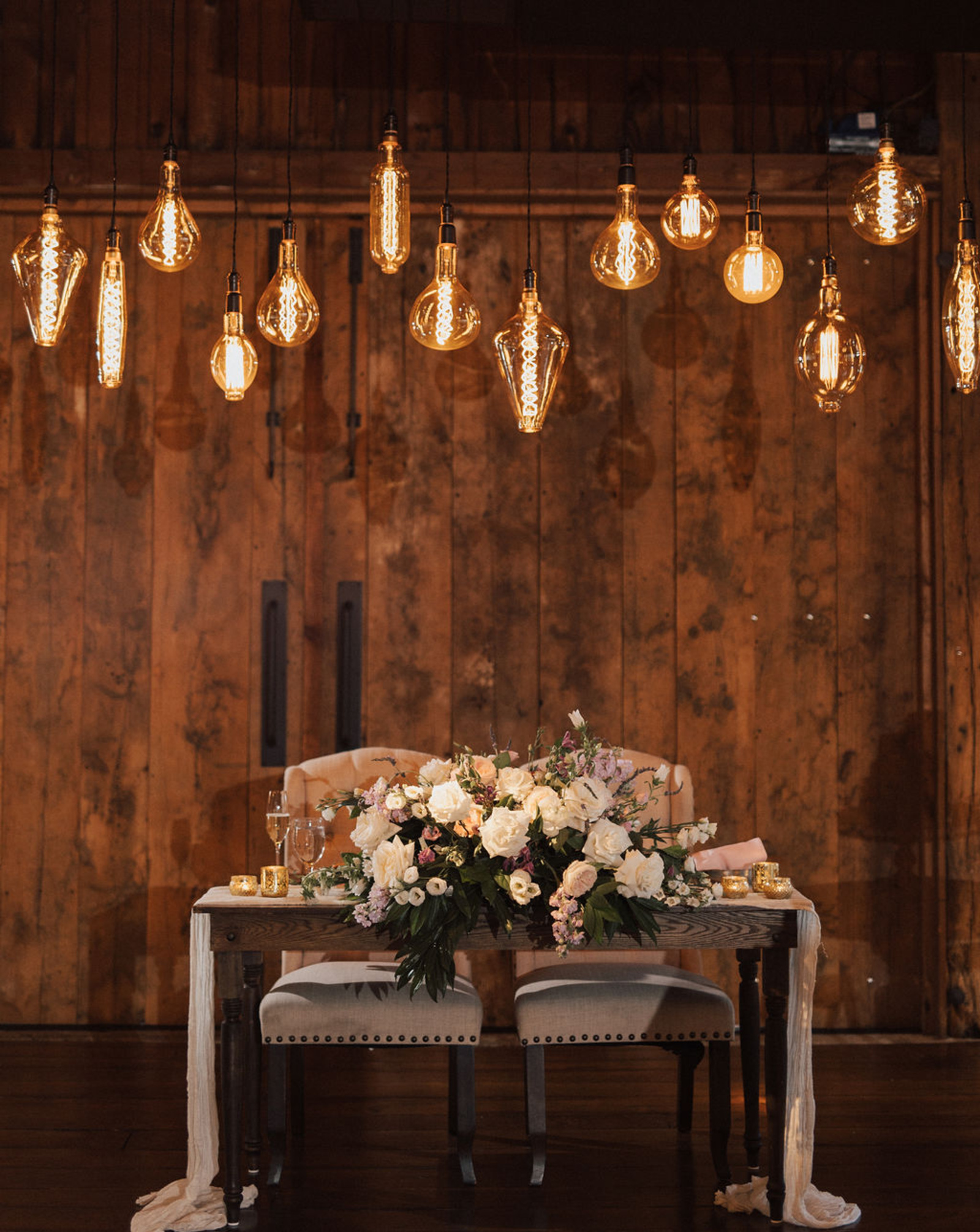 Romantic sweetheart table draped in white linens and white rose floral bouquet under 14 string Edison lightbulb lights