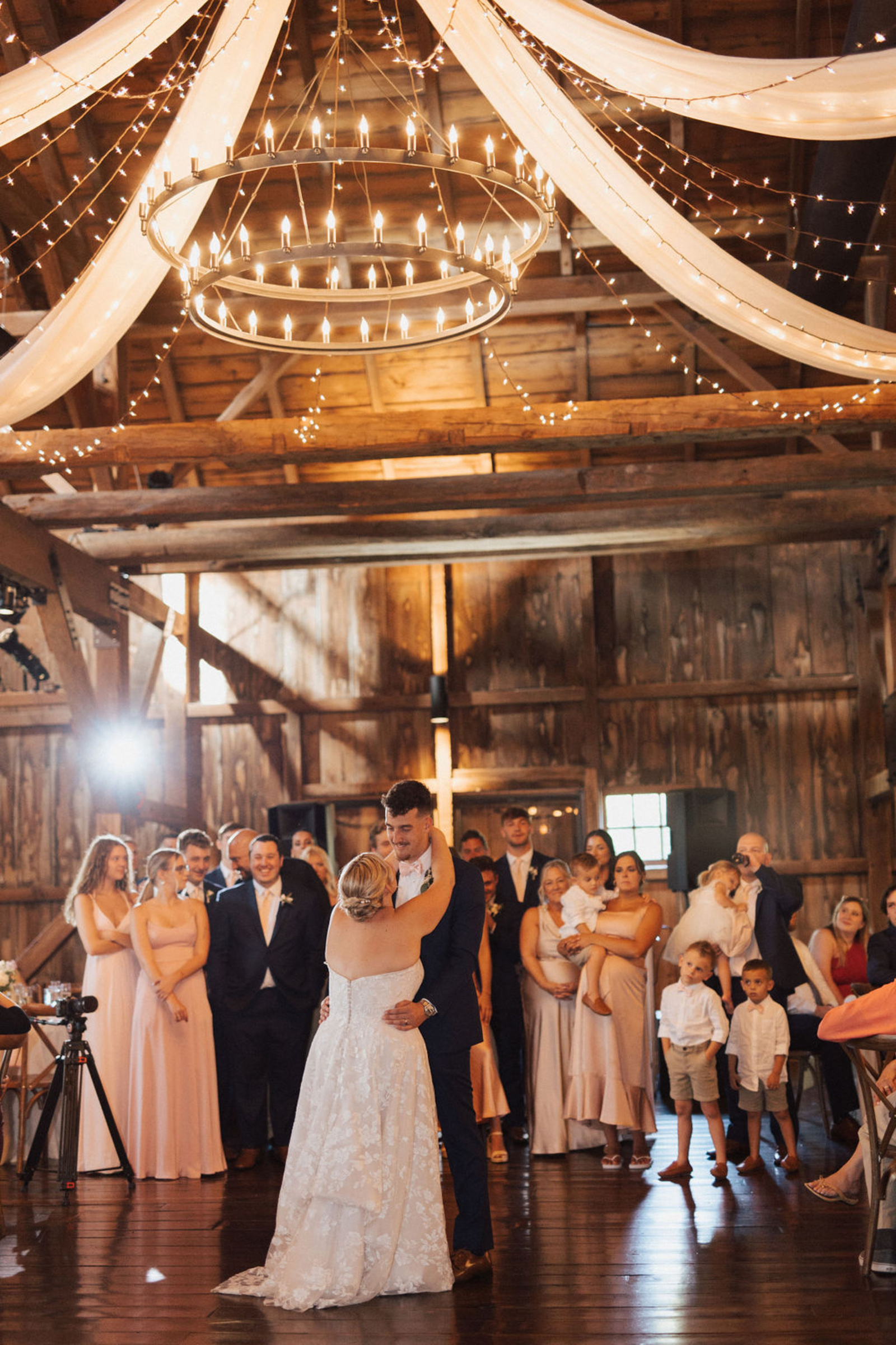 Central PA bride and groom dancing under a two-tiered chandelier in a rustic wood venue surrounded by family and friends