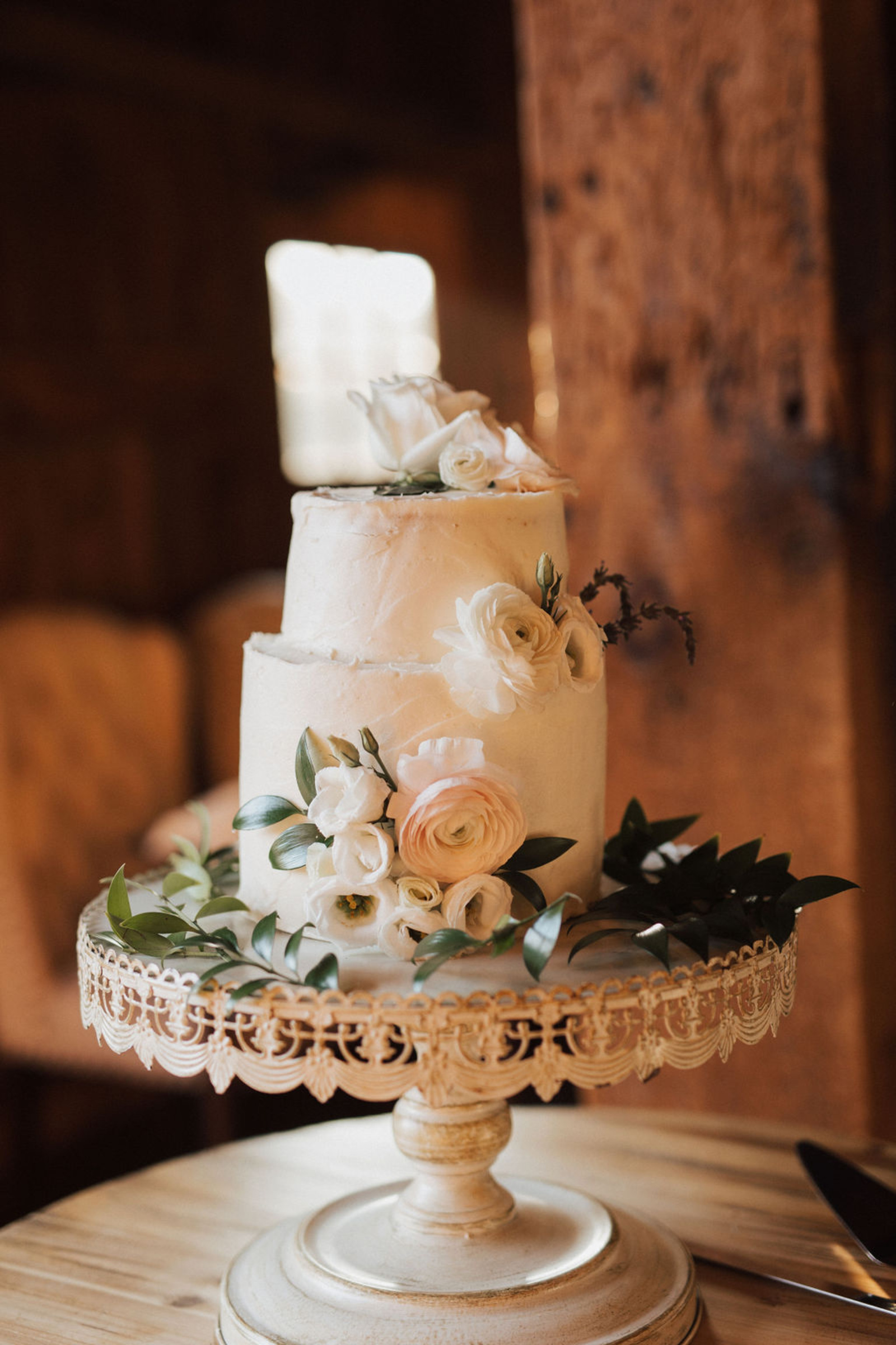 Simple white two-tiered wedding cake adorned with white and pink flowers and greens on top of an antique white cake stand