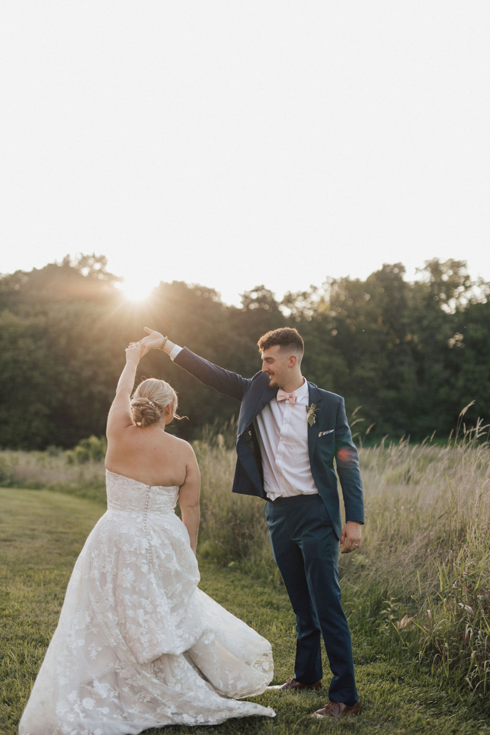 Groom wearing a navy suit and pink bow tie spinning bride in a strapless lace dress in a Central PA field