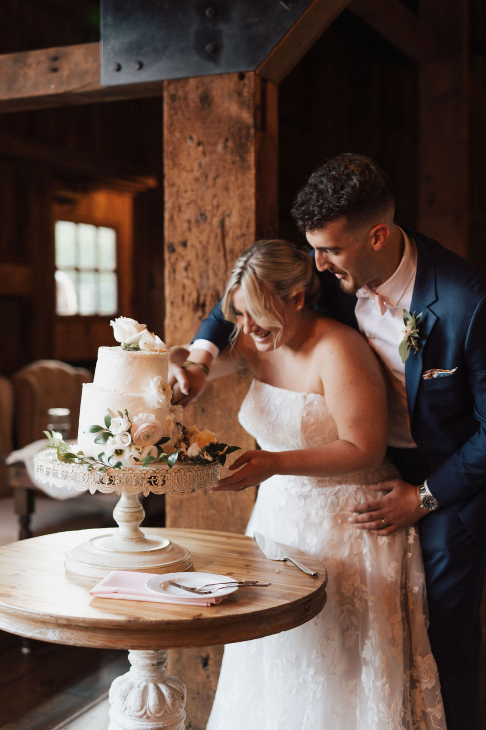 Central PA bride and groom smiling while cutting a simple  white two-tiered wedding cake adorned with white and pink flowers and greens on top of an antique white cake stand