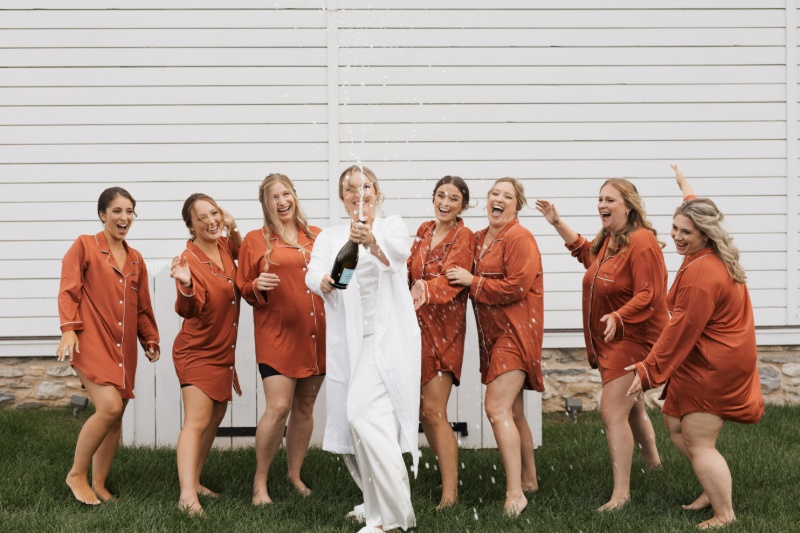 Bride popping champagne with her bridesmaids in rust-colored getting-ready outfits before a fall wedding at St. Michael’s Vineyard.