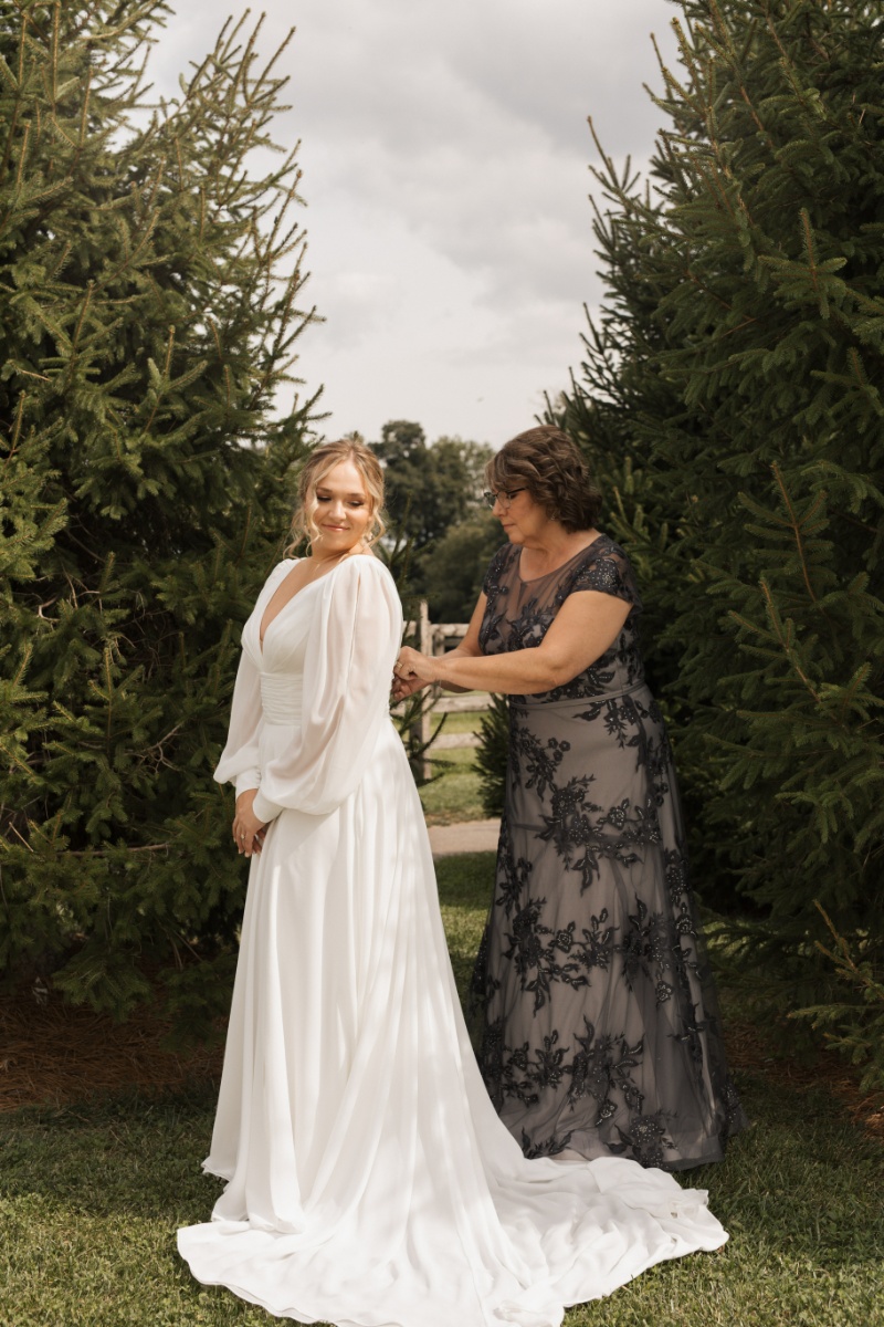 Bride in a long-sleeve wedding gown getting help with her dress outdoors among evergreen trees before her St. Michael’s Vineyard wedding.