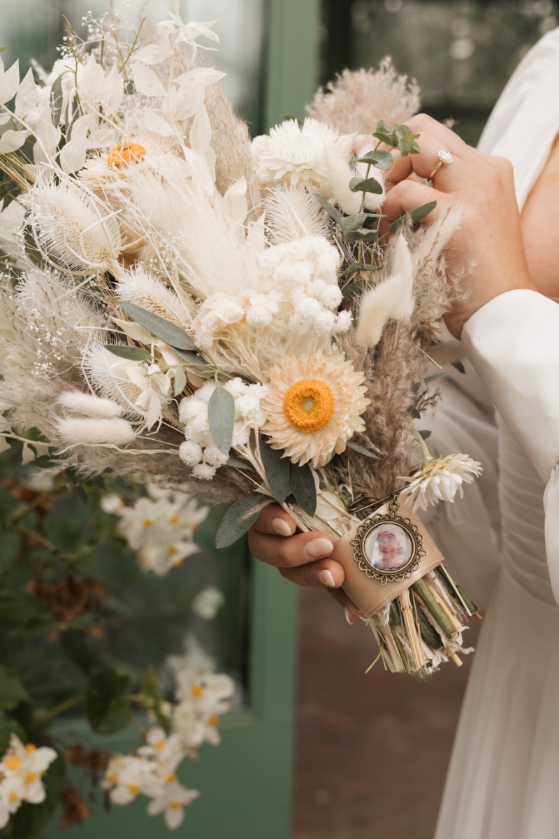 Bride holding a textured fall wedding bouquet with cream dried florals, eucalyptus, soft grasses, and a sentimental photo charm.