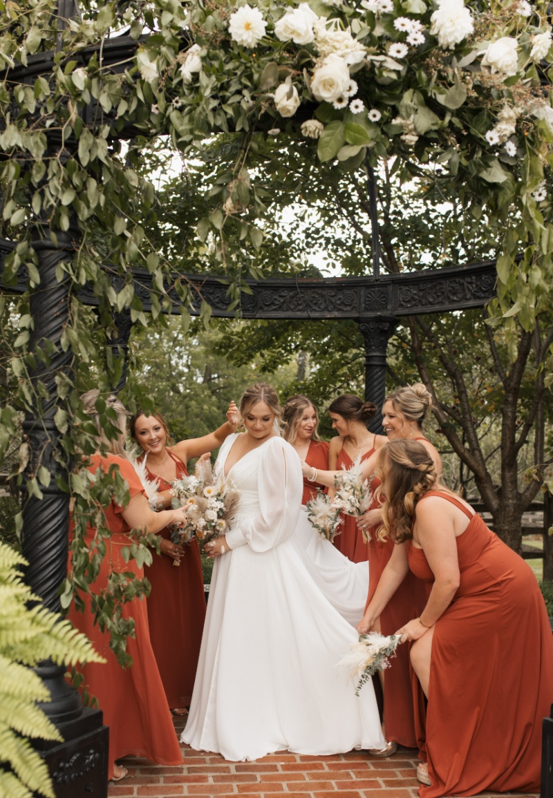 Bride surrounded by bridesmaids in rust-colored dresses beneath a flower-covered black garden gazebo at St. Michael’s Vineyard.