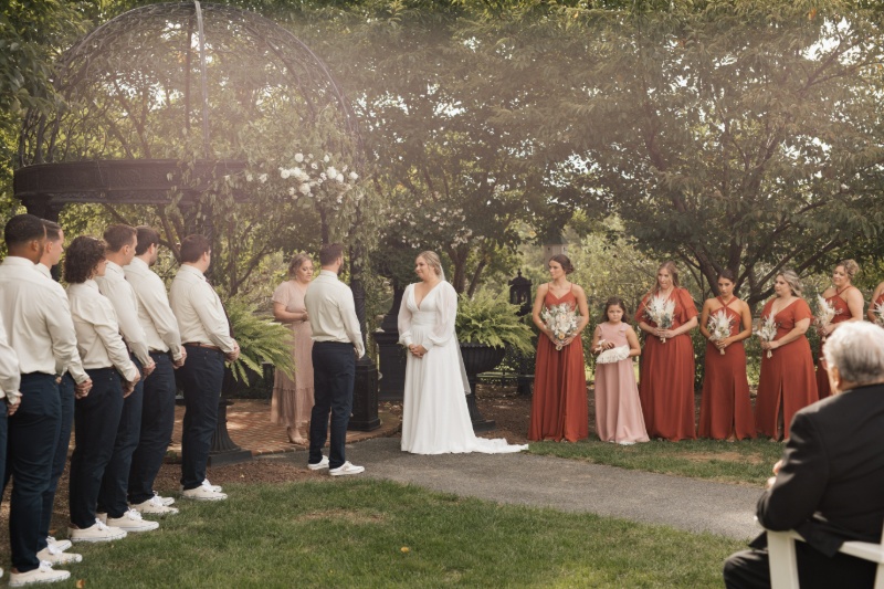 Bride and groom exchanging vows during an outdoor fall wedding ceremony at St. Michael’s Vineyard, surrounded by bridesmaids in rust dresses and groomsmen in neutral shirts.