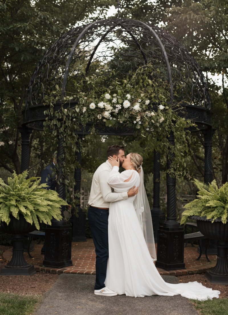 Newlyweds kissing beneath a black garden gazebo decorated with white flowers and greenery after their fall wedding ceremony at St. Michael’s Vineyard.