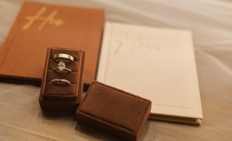 Engagement ring and wedding bands displayed in a brown velvet ring box beside personalized wedding vow books.