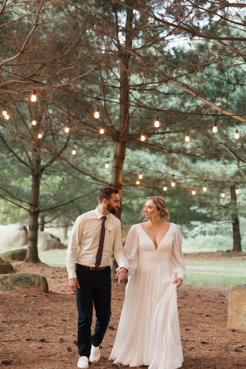Bride and groom walking hand in hand beneath string lights among the trees during their fall wedding at St. Michael’s Vineyard.