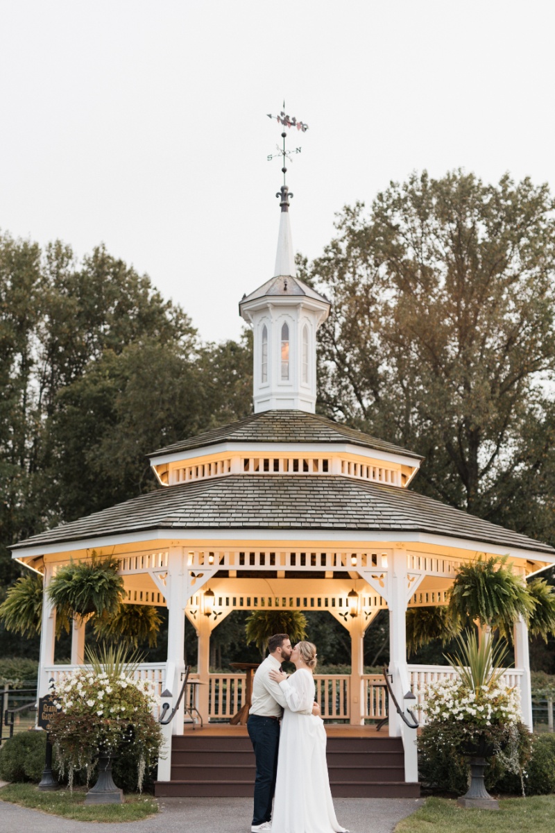 Bride and groom kissing in front of a white gazebo decorated with flowers at St. Michael’s Vineyard.