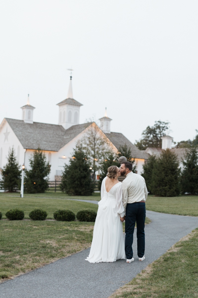Bride and groom walking hand in hand toward the white barn at St. Michael’s Vineyard after their fall wedding ceremony.