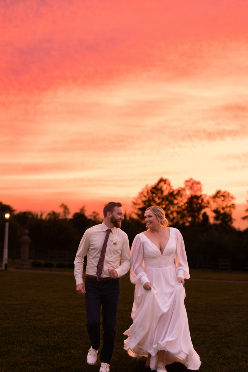 Bride and groom running together beneath a dramatic orange and pink sunset during their fall wedding at St. Michael’s Vineyard.