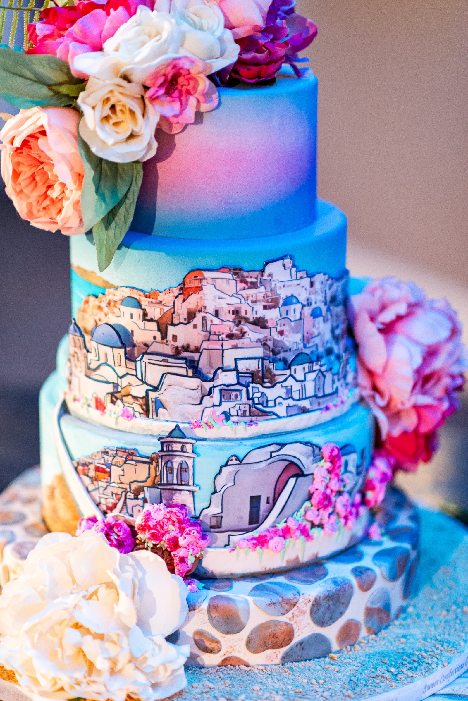 A three-tier wedding cake decorated with hand-painted scenes of a Mediterranean village, featuring white and blue domed buildings resembling Santorini. The cake is topped with vibrant pink, peach, and white flowers, accented with greenery. The base tier is designed with a stone-like pattern, and the cake is displayed on a sandy surface surrounded by small candles.