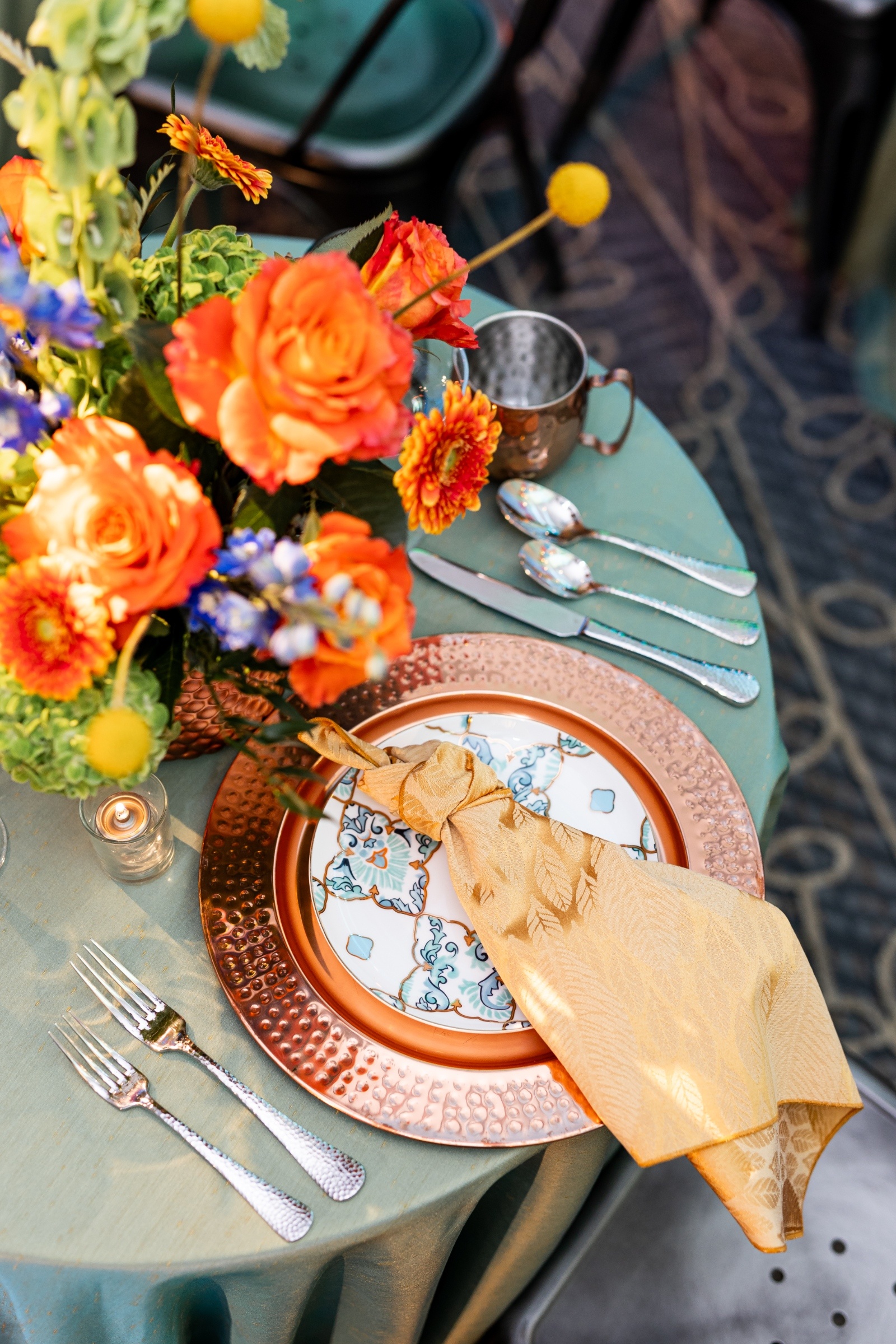 Wedding tablescape featuring copper charger plates, yellow napkins, and a vibrant floral centerpiece with verona orange orange roses and blue blooms.