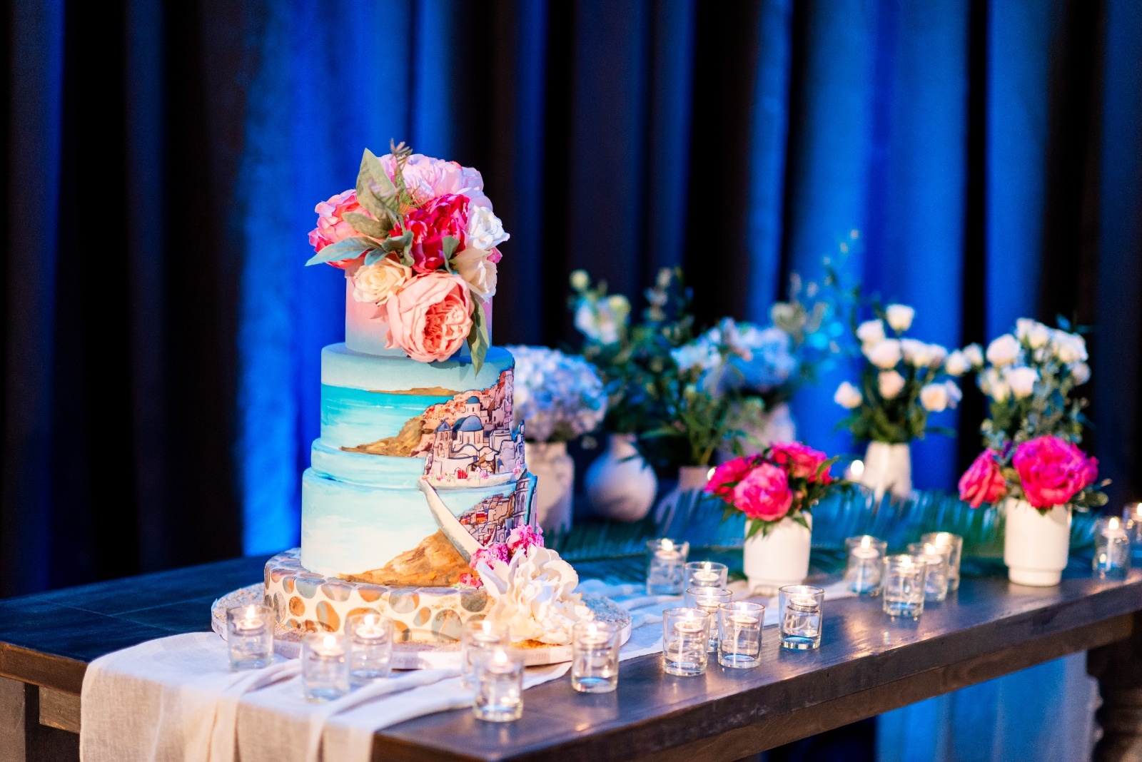 A vibrant, hand-painted wedding cake inspired by the Greek island of Santorini, displayed on a rustic wooden table with glowing candles and floral accents. The cake features ocean blues, coastal scenery, and pink floral details under moody blue lighting.