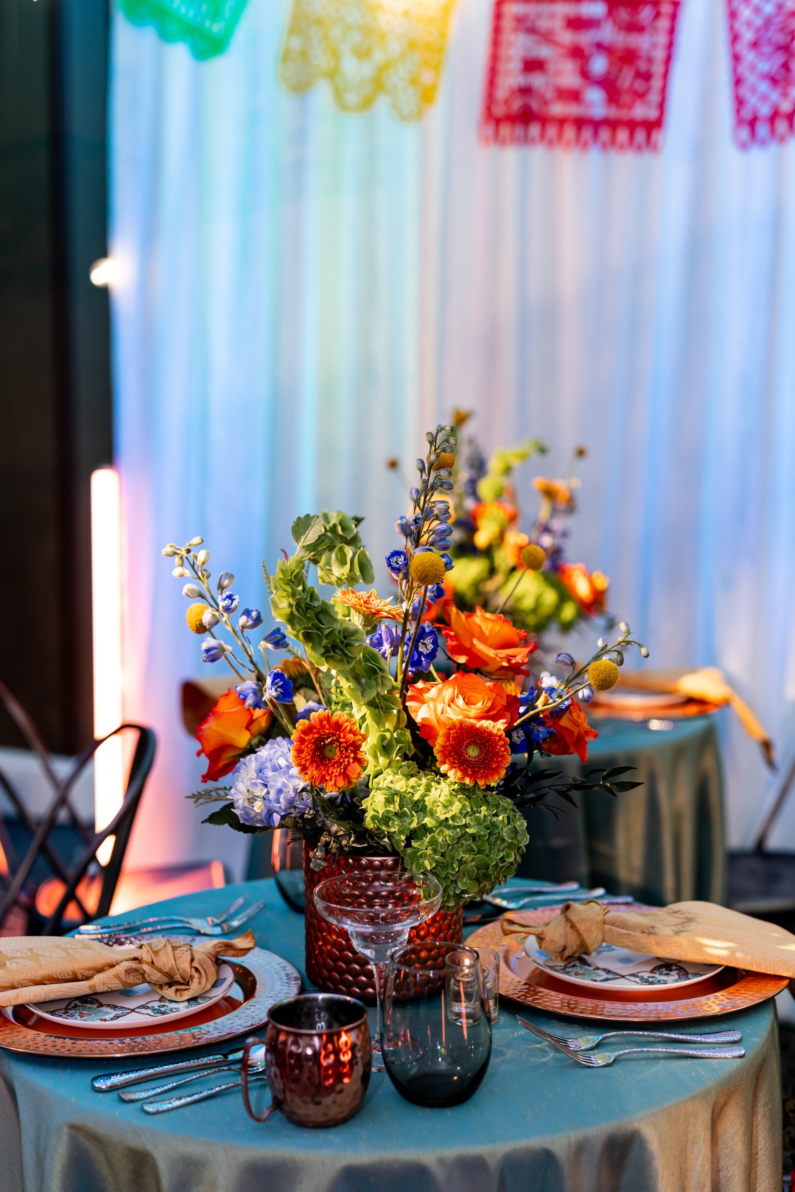 Reception table with bright verona sunset orange and blue florals beneath papel picado banners, styled with copper mugs and teal linens for a festive celebration.