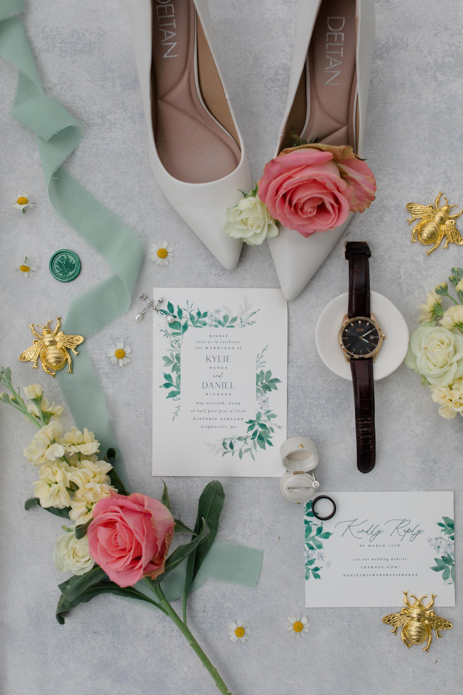Wedding Flat Lay with Shoes, Invitation Suite, and Florals Wedding flat lay featuring bridal shoes, invitation suite, ribbon, watch, and florals styled on a light textured background.