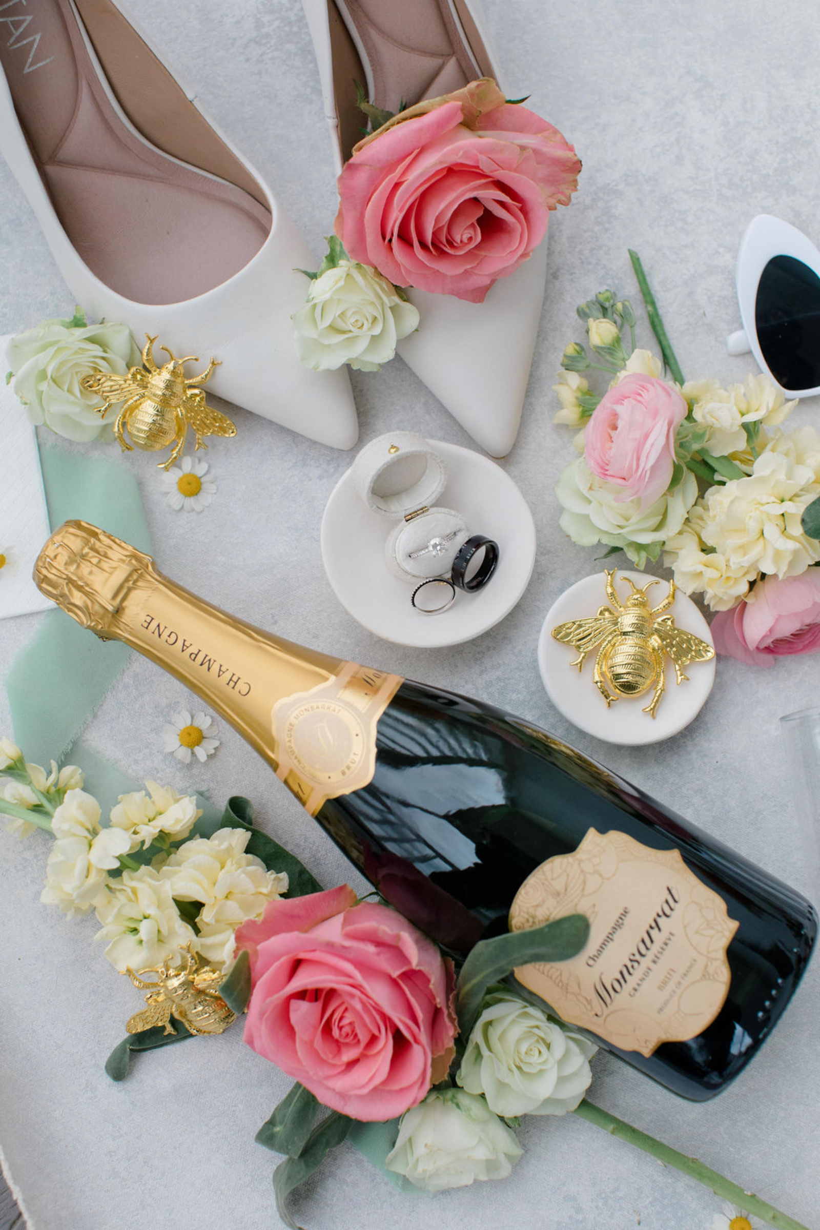 A beautifully styled wedding flat lay featuring a bottle of champagne, blush and ivory roses, gold bee accents, and the couple’s rings — captured at Historic Ashland.