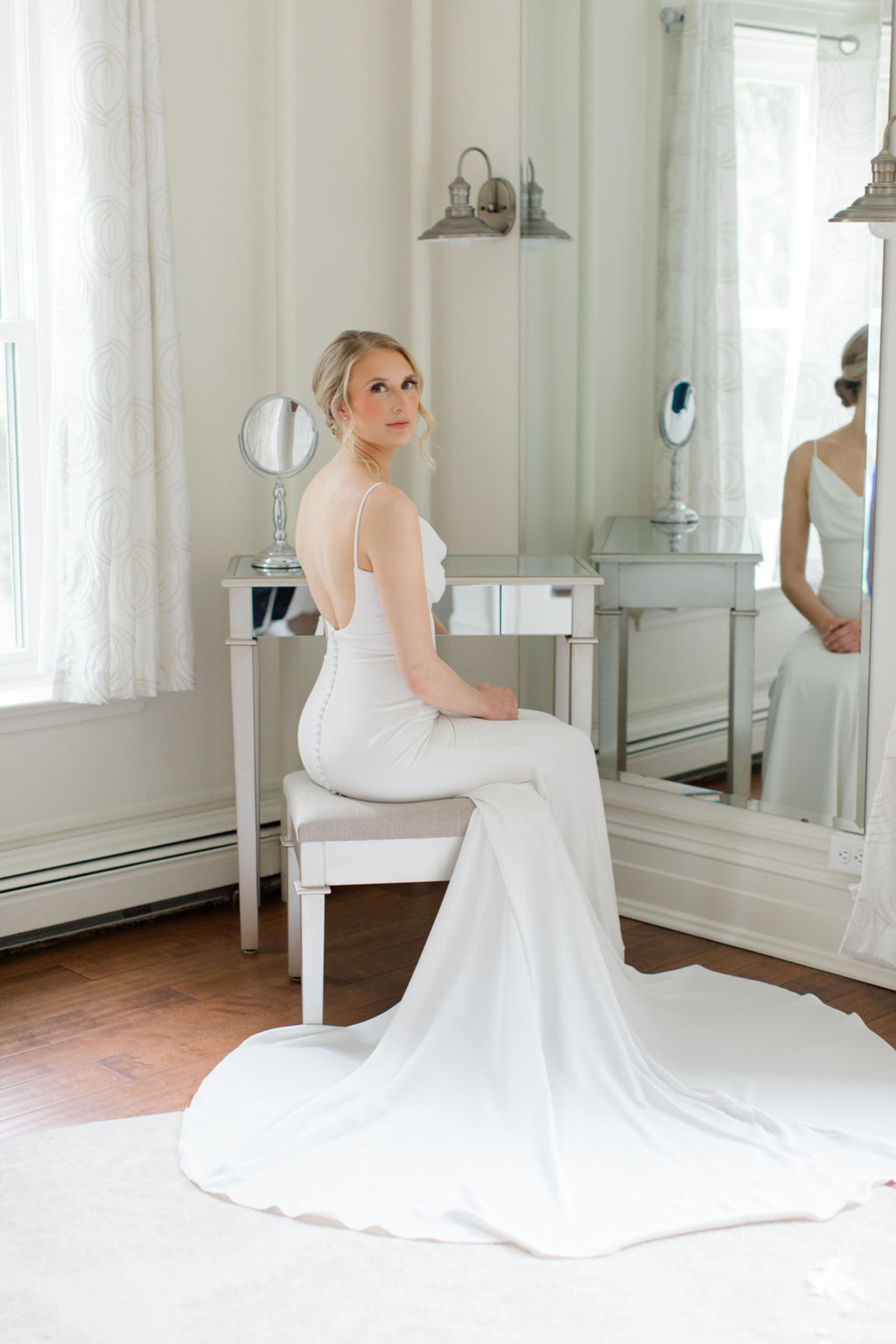 The bride poses elegantly in a fitted white gown with button-back detailing while sitting at a mirrored vanity in the bridal suite at Historic Ashland.