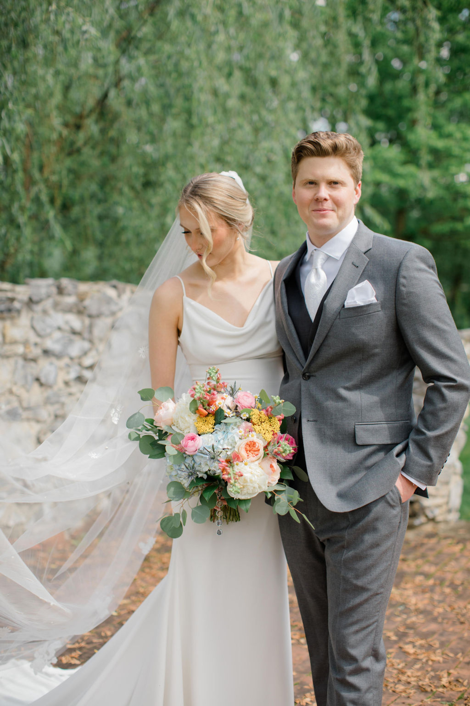 The bride and groom pose for a romantic portrait on the grounds of Historic Ashland, with the bride holding a colorful spring bouquet and her veil flowing softly behind her.