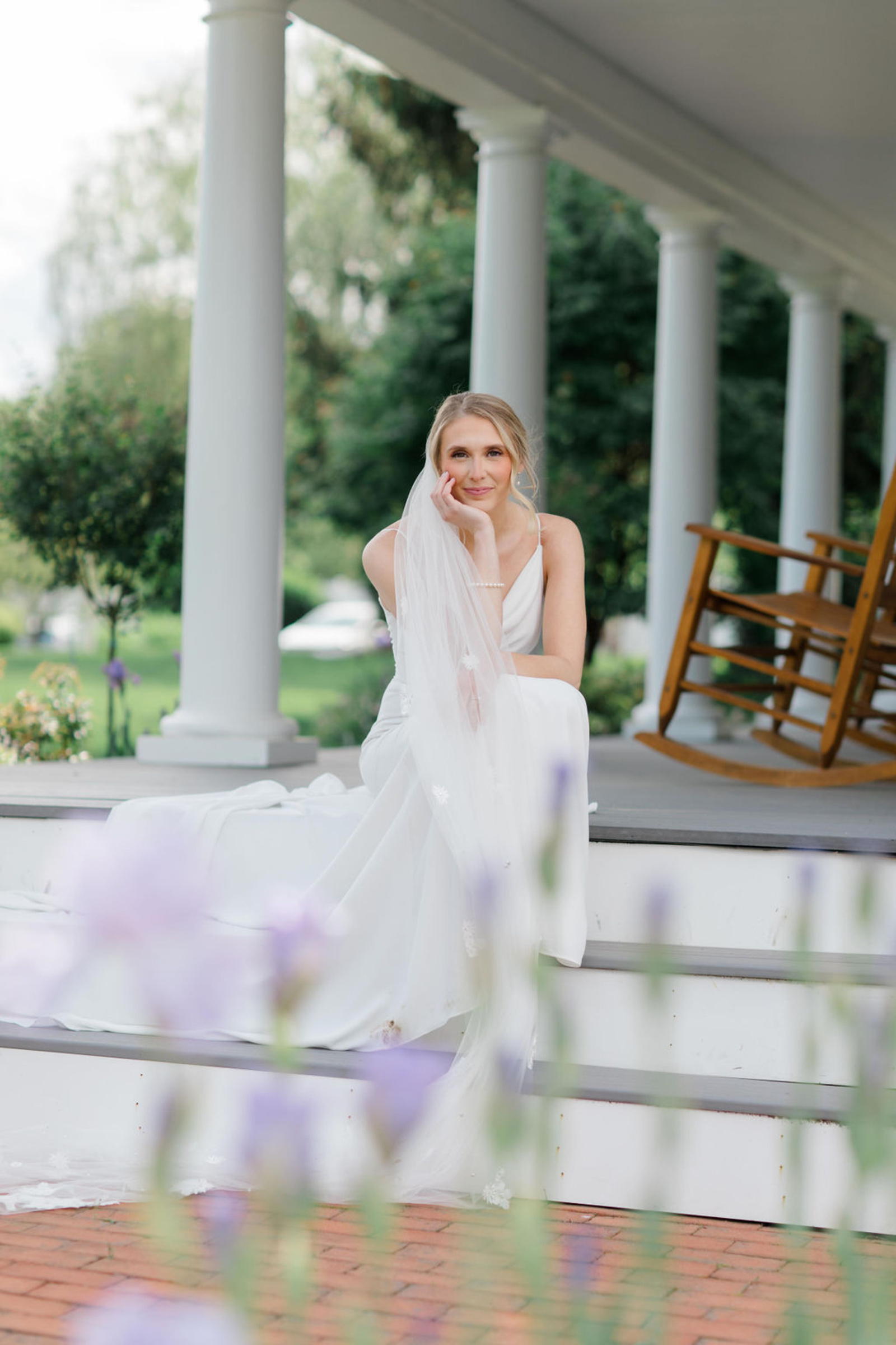 The bride sits gracefully on the front porch steps of Historic Ashland, resting her chin on her hand with a soft smile surrounded by fresh greenery and soft purple blooms.