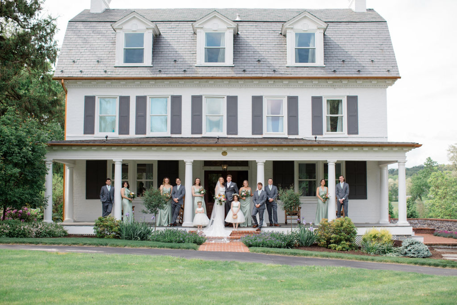 The bride and groom stand with their wedding party on the grand front porch of Historic Ashland, surrounded by lush gardens and classic white columns.