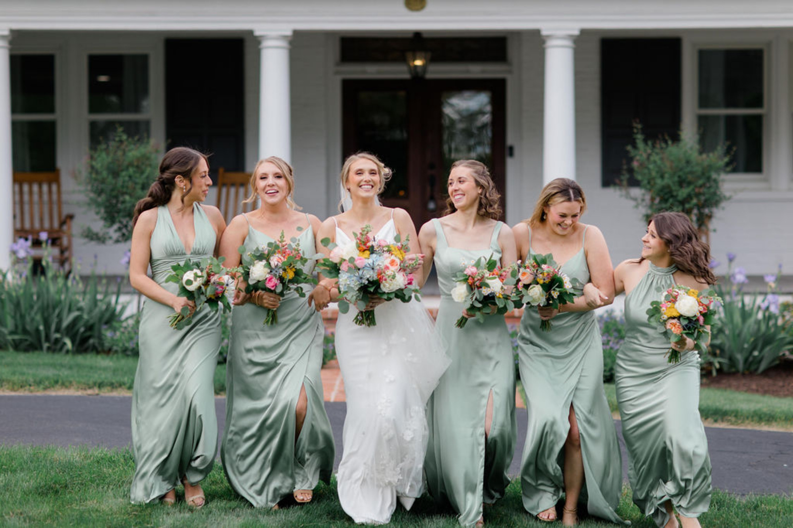 The bride and her bridesmaids, dressed in mismatched sage green gowns, laugh and walk together across the lawn at Historic Ashland holding colorful spring bouquets.