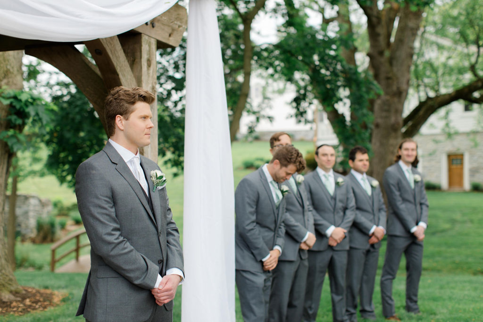 Groom and groomsmen in gray suits stand at the altar beneath a wooden arbor during an outdoor ceremony at Historic Ashland.