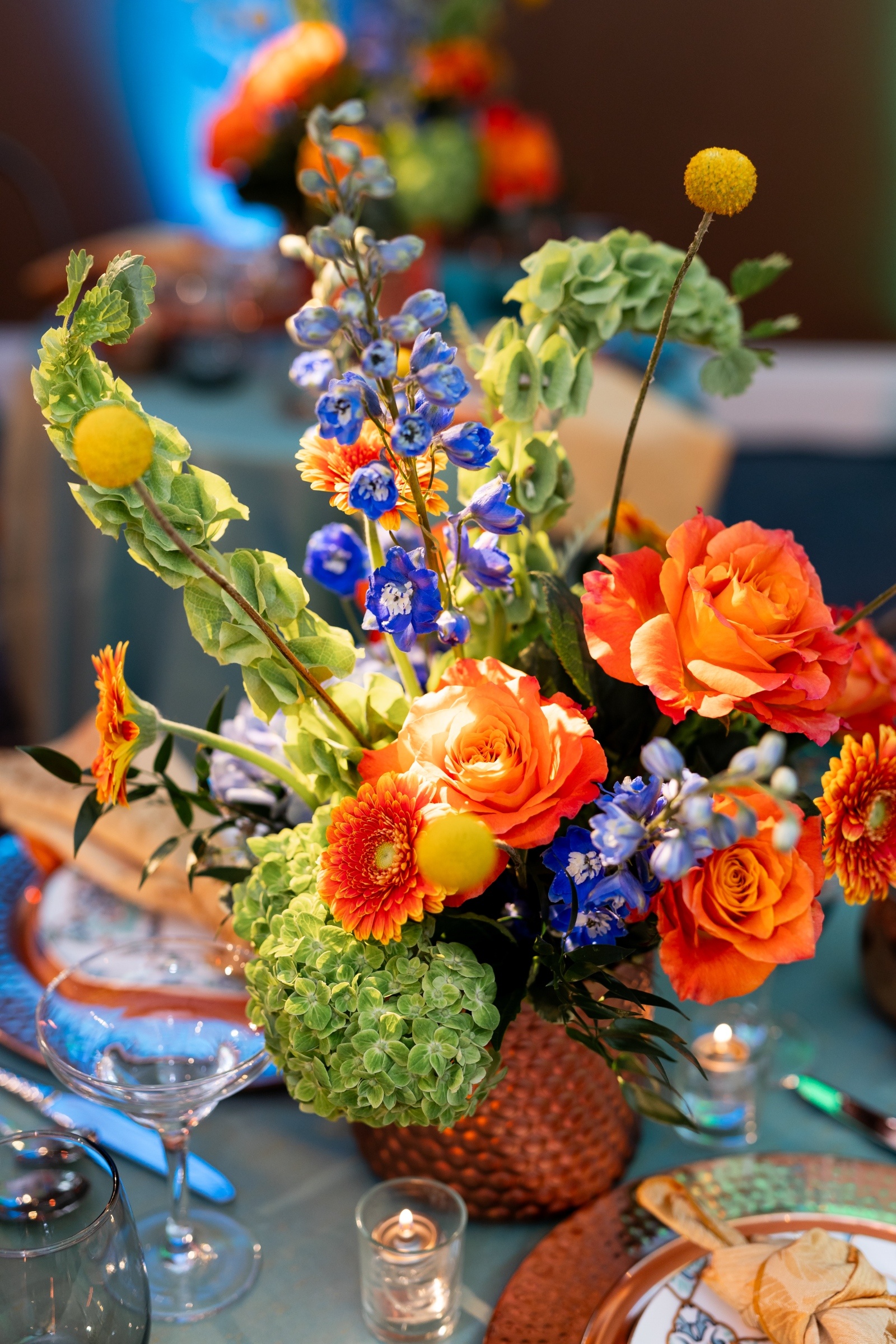 Close-up of a wedding centerpiece with verona sunset orange roses, green hydrangeas, yellow billy balls, and blue delphinium in a hammered copper vase.