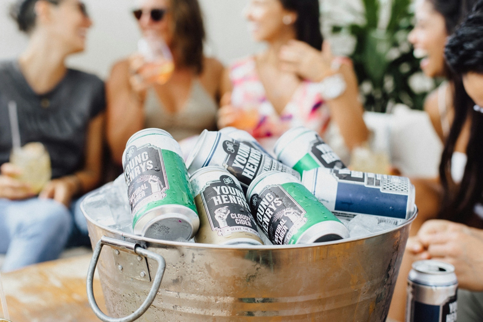 Close-up of a metal bucket filled with cold canned drinks as a group of friends laugh and enjoy cocktails together during a sunny bachelorette weekend celebration.