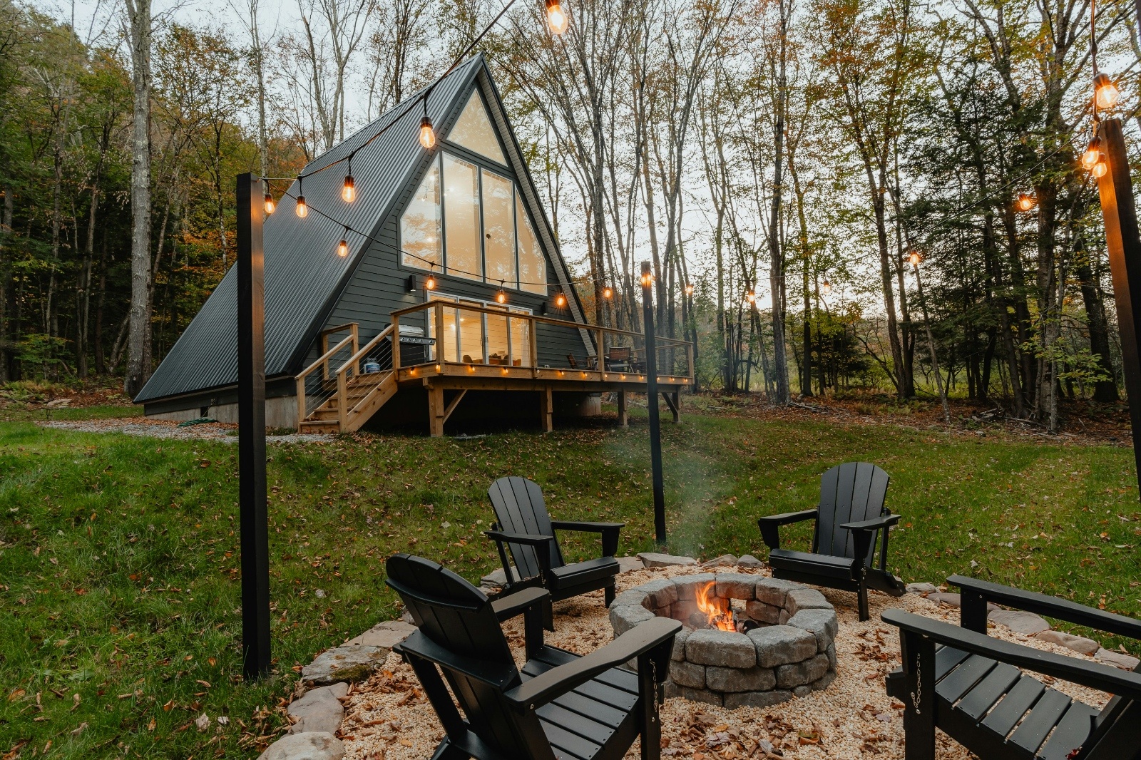 Cozy modern A-frame cabin with large glass windows and string lights glowing at dusk, surrounded by autumn trees. Four black Adirondack chairs circle a stone fire pit in the grassy backyard, creating a warm and inviting outdoor retreat.