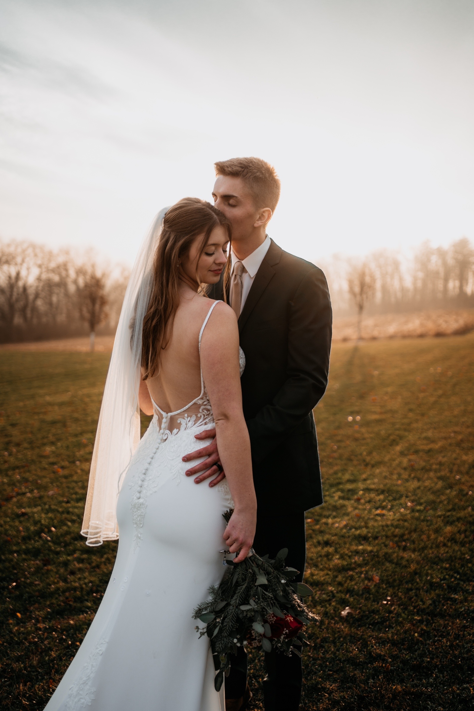 Groom kisses bride’s forehead at sunset as she holds a greenery bouquet, captured in warm golden light by Téa Ceresini Photography.