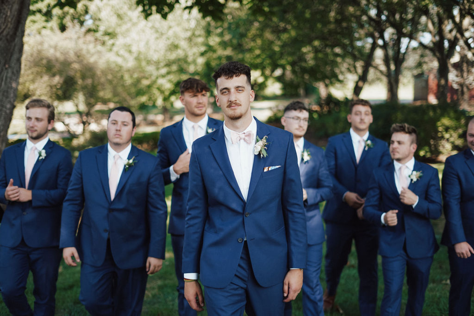 Central PA groom in a navy blue suit walking towards the camera with a straight face with 7 groomsmen in navy suits walking behind him